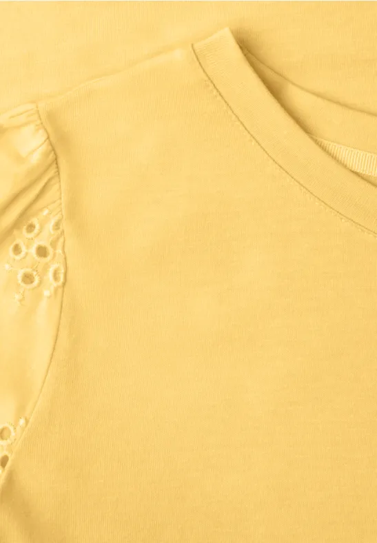 Nahaufnahme eines gelben T-Shirts mit Rundhalsausschnitt und filigranen Lochstickereien an der Schulter.