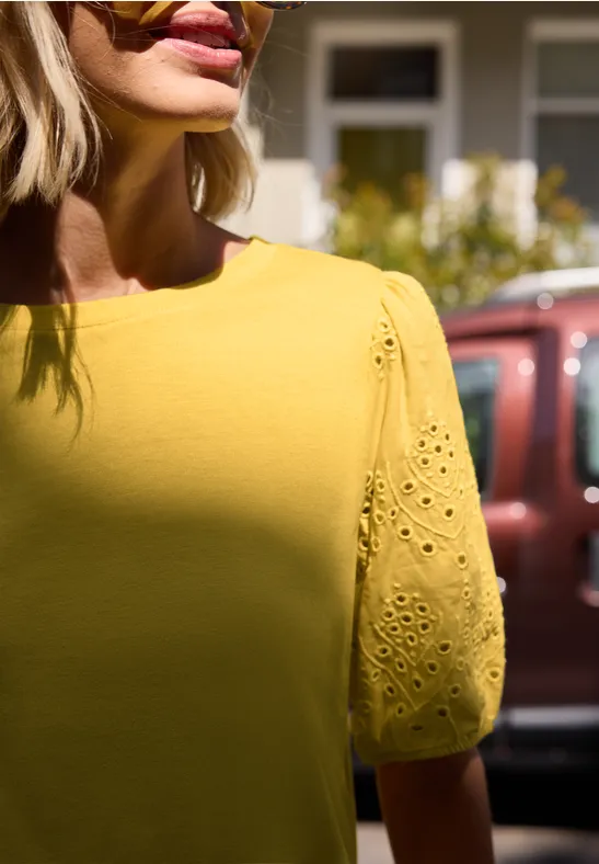 Frau trägt gelbes Shirt mit bestickten Kurzärmeln, Nahaufnahme, unscharfer Hintergrund.