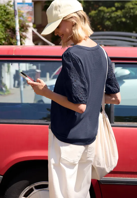 Frau in Kapuzenpulli, weißer Hose und Mütze blickt auf ihr Handy neben rotem Auto.