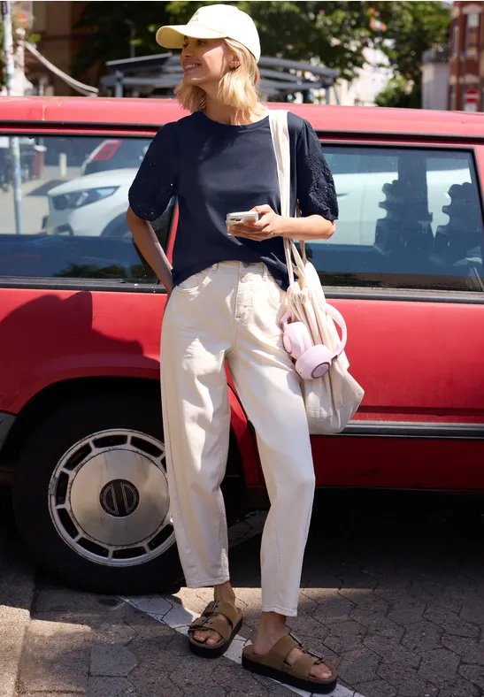 Person in T-shirt, weiten Hosen und Sandalen steht vor rotem Auto, hält Smartphone und hat Tasche mit Kuscheltier.