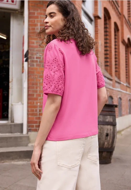 Person in pinker Bluse mit Lochstickerei-Ärmeln und hellen Hosen vor rotem Backsteingebäude.