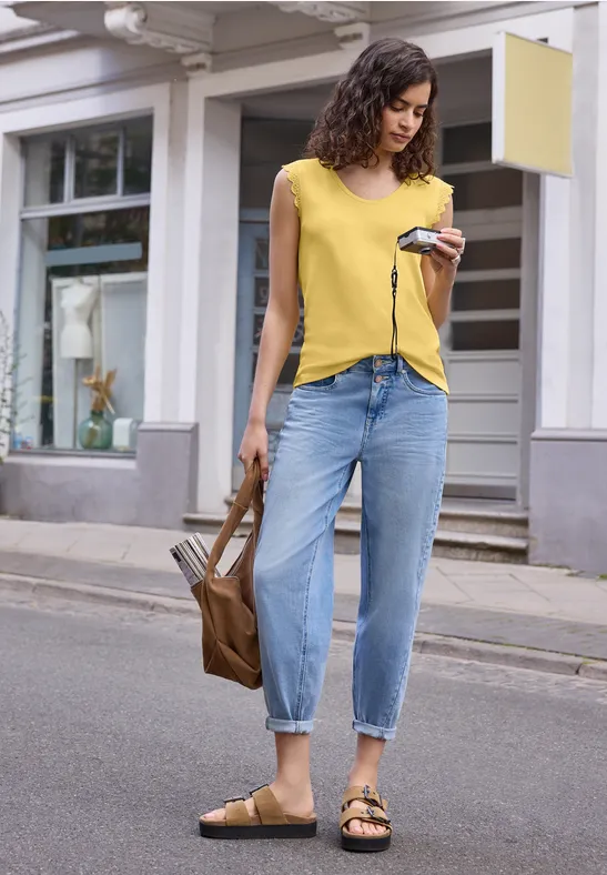 Frau in gelbem T-Shirt, blauen Jeans und Sandalen, hält Smartphone und braune Tasche auf der Straße.