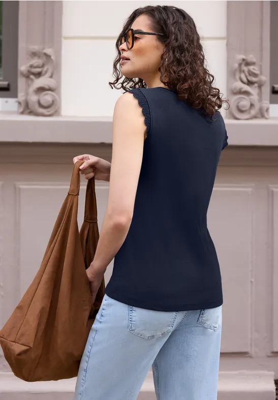 Frau mit lockigem Haar in ärmelloser schwarzer Bluse, hellen Jeans, trägt eine braune Tasche vor rosa Hintergrund.