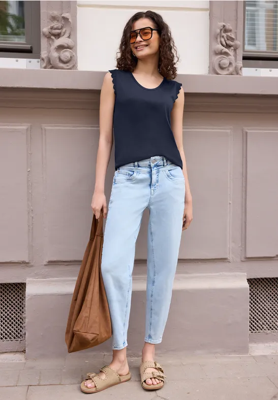 Eine Person steht vor einer Wand mit Ornamenten, trägt hellblaue Jeans, dunkelblaues Top, Sandalen und Tasche.