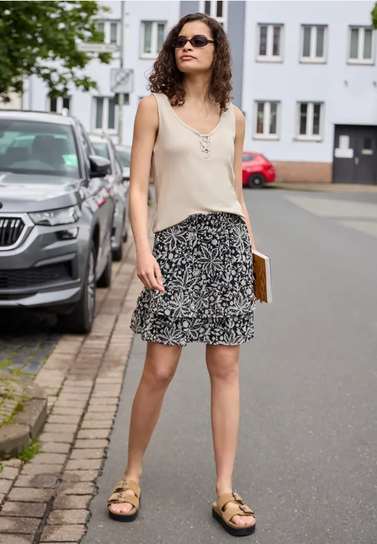 Frau in beigem Oberteil, gemustertem Rock und Sandalen hält ein Notizbuch auf dem Gehweg vor Häusern.