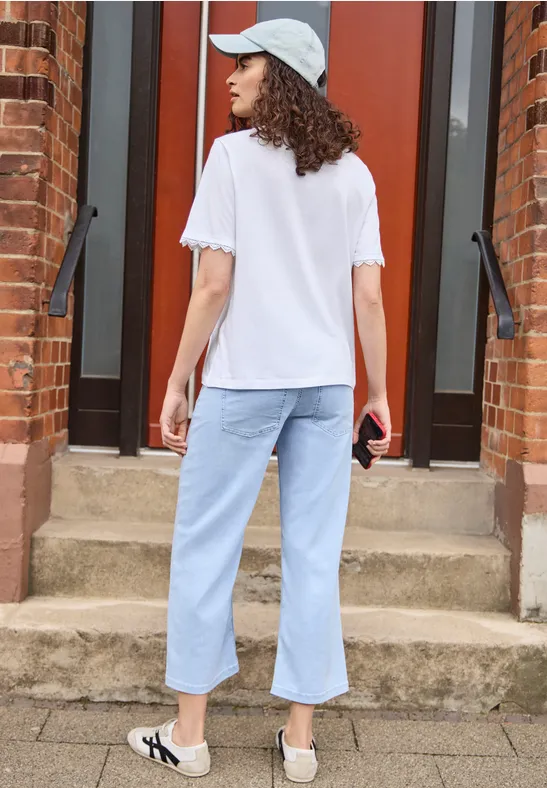 Frau von hinten in weißem T-Shirt, hellen Jeans und Mütze steht vor einer Backsteintür auf Stufen.