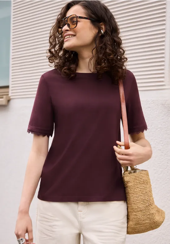 Frau mit hüftlanger, lockiger Frisur trägt bordeauxfarbenes T-Shirt, helle Hose und Rattantasche.