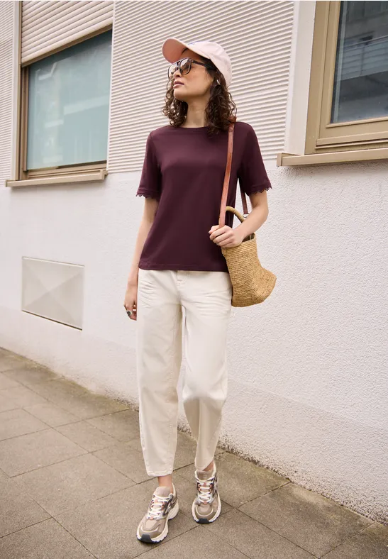 Eine junge Frau in lila T-Shirt, weißen Hosen und Cap mit Strohtasche steht auf dem Bürgersteig.