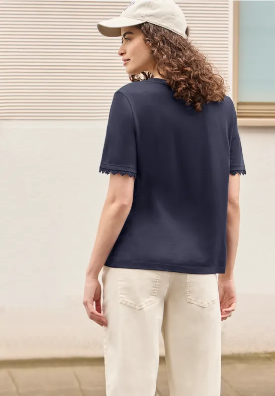 Persoon gezien van achteren met donkerblauw T-shirt, beige broek en witte pet voor een lichte muur.