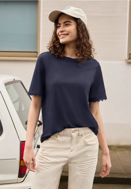 Een vrouw staat voor een auto, draagt een blauw T-shirt, een lichte broek en een witte pet.