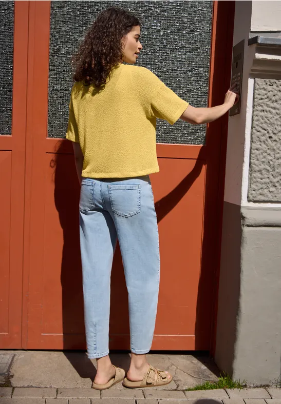 Eine Frau mit gelbem T-Shirt, blauen Jeans und Sandalen steht vor einer roten Tür und blickt zur Seite.