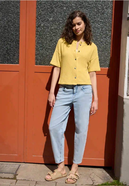 Person in gelbem Hemd und blauer Jeans vor einer rötlichen Tür.