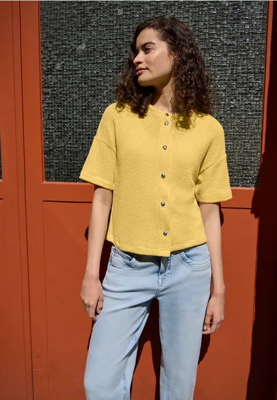 Eine Frau in gelbem Kurzarmshirt und blauen Jeans steht vor einer roten Tür.