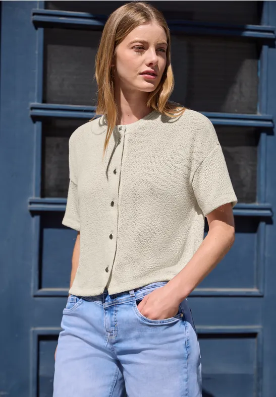 Eine Frau trägt eine kurzärmelige, helle Bluse und Jeans und steht vor einer blauen Tür.