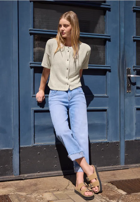 Frau lehnt vor einer blauen Tür, trägt helle Bluse, blaue Jeans und Sandalen.