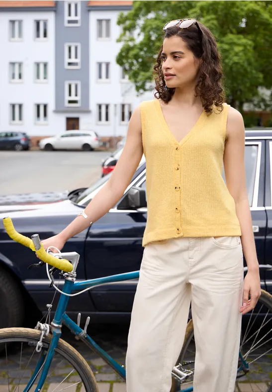 Person mit gelbem Top und weißer Hose steht mit Fahrrad vor einem Wohnhaus.