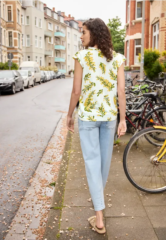 Een vrouw met krullend haar in een wit topje met groen patroon en lichte jeans loopt op het trottoir.