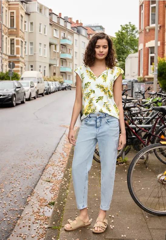 Een vrouw in een bloes met bloemen, lichte jeans en sandalen staat op een stadsstraat naast geparkeerde fietsen.