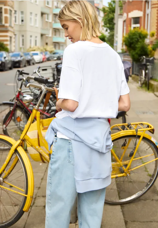 Eine Person in weißem T-Shirt und Jeans steht neben einem gelben Fahrrad in einer Stadtstraße.