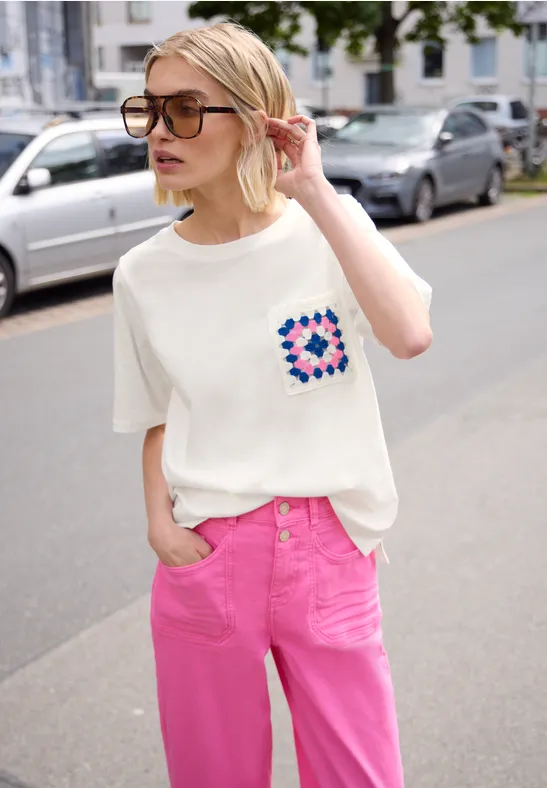 Person auf Straße vor parkenden Autos trägt weißes T-Shirt mit buntem Taschenmuster und pinke Hose.