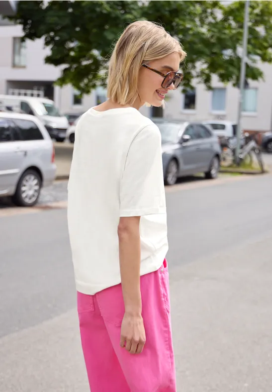 Frau mit weißem T-Shirt und pinker Hose steht auf einer Straße, Autos und Fahrräder im Hintergrund.