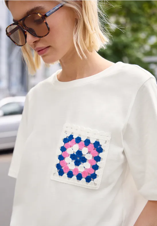 Person trägt weißes T-Shirt mit gehäkelter Tasche in buntem Muster (blau, rosa, weiß).