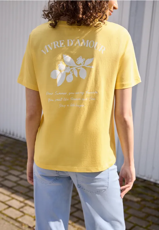 Eine Person trägt ein gelbes T-Shirt mit dem Aufdruck ‚VIVRE D'AMOUR‘ und einem weißen Blumenmotiv.