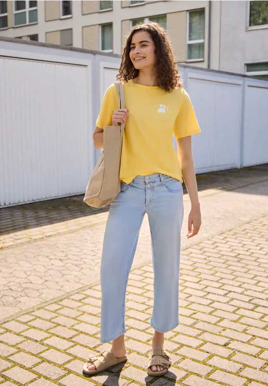 Frau in gelbem T-Shirt, hellblauen Jeans und Sandalen hält eine beige Tasche und steht auf gepflastertem Hof.