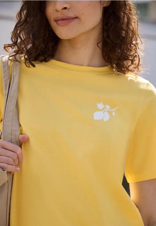 Eine Frau trägt ein gelbes T-Shirt mit weißem Blumen-Logo und Buchstaben.
