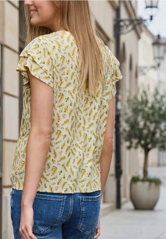 Vrouw in een geel, motiefblouse en spijkerbroek, zijwaarts staand op een stadsstoep.
