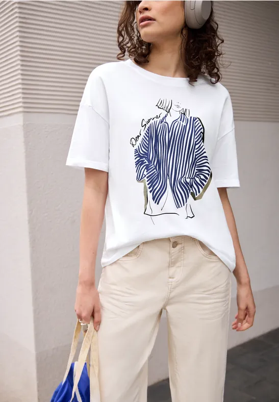 Persoon in beige broek en wit T-shirt met blauw lijnenschets-print, draagt koptelefoon en rieten tas.