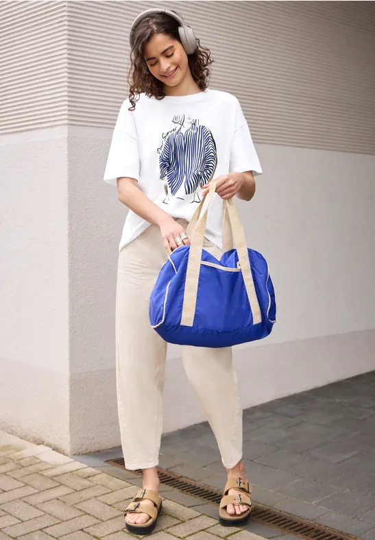 Jonge vrouw met koptelefoon, wit T-shirt, beige broek en blauwe reistas