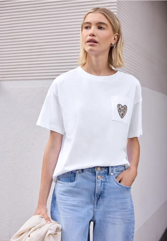 Frau in weißem T-Shirt mit Herz-Applikation und blauen Jeans, eine Hand in der Tasche, vor moderner Wand