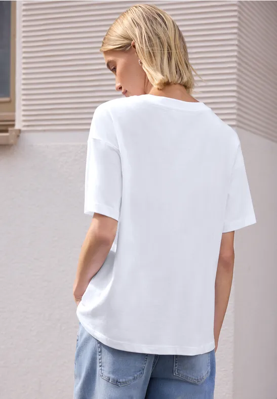 Eine Frau mit blonden Haaren steht seitlich-von hinten vor einer hellen Wand und trägt ein weißes T-Shirt.