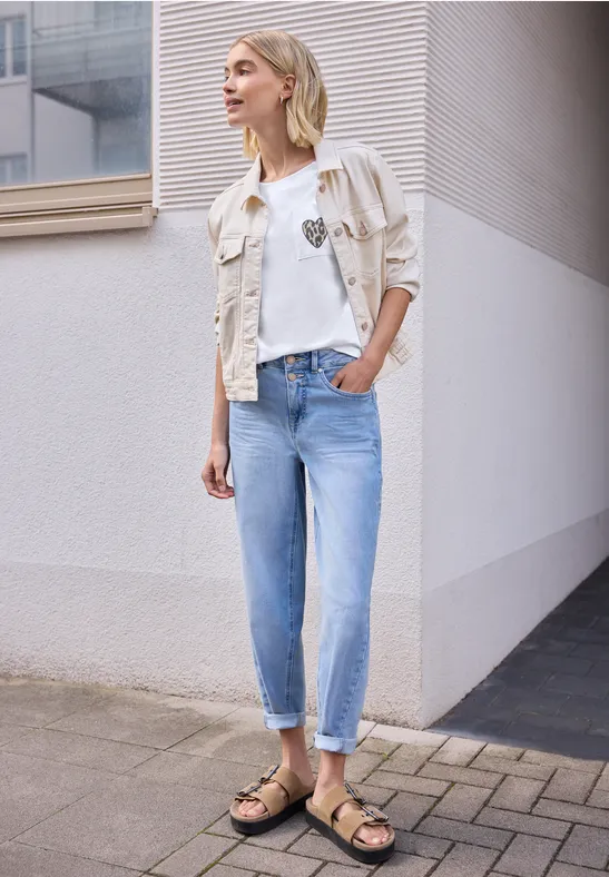 Frau in heller Jeans, weißem Shirt und Jeansjacke mit Sandalen steht vor modernem Gebäude.