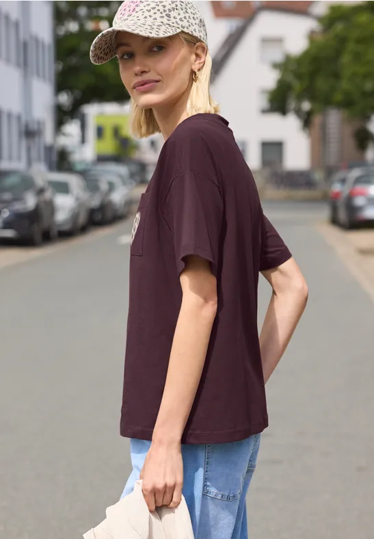 Person im lila T-Shirt und Jeans steht seitlich auf einer ruhigen Wohnstraße mit geparkten Autos.