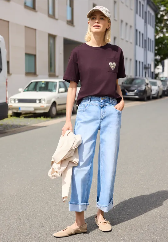 Person in lila T-Shirt mit Herz-Patch, weiten, hochgekrempelten Jeans und Loafers, hält Jacke auf Straße.