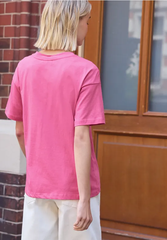 Frau im pinken T-Shirt und hellen Hosen, von hinten vor einem Haus stehend.