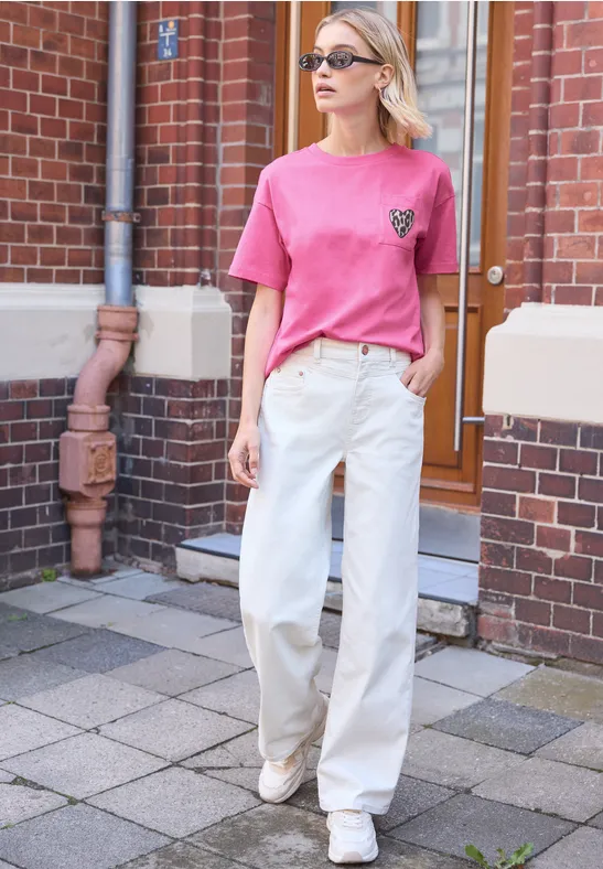 Frau vor Backsteinhaus trägt pinkes T-Shirt und weiße weite Hose, lässiger Streetstyle.