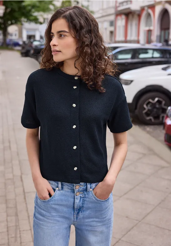 Frau in schwarzem Kurzarmpullover mit Perlenknöpfen und hellblauen Jeans steht auf dem Gehweg vor parkenden Autos.