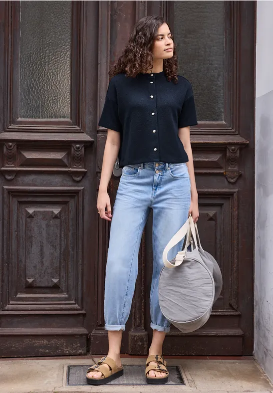 Junge Frau in schwarzer Bluse, hellen Cropped-Jeans und Sandalen mit grauer Tasche vor einer Holztür.