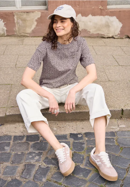Persoon zit ontspannen op een laag muurtje, met shirt, lichte broek en sneakers.