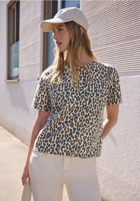 Frau im Leoparden-T-Shirt, in weißer Hose und Basecap, steht vor sonniger Hauswand