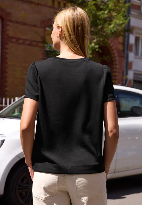 Eine blonde Frau steht von hinten in schwarzem T-Shirt und beiger Hose vor einem weißen Auto.
