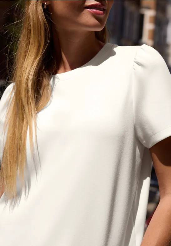 Une femme sous le soleil porte un chemisier blanc et arbore de longs cheveux blonds.