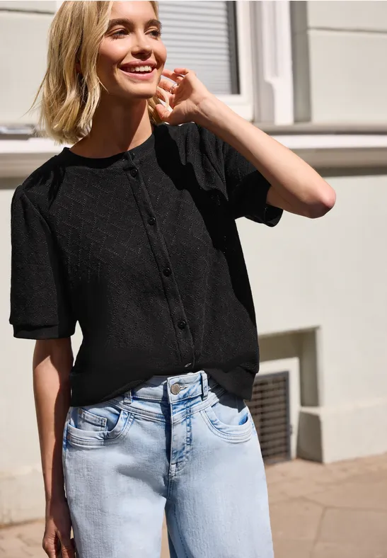Frau trägt schwarzes, knöpfbares Kurzarmshirt und blaue Jeans vor einer Hausfassade.