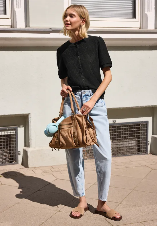 Frau mit schwarzem Oberteil und hellen Jeans steht vor einer Hauswand und hält eine große braune Tasche.