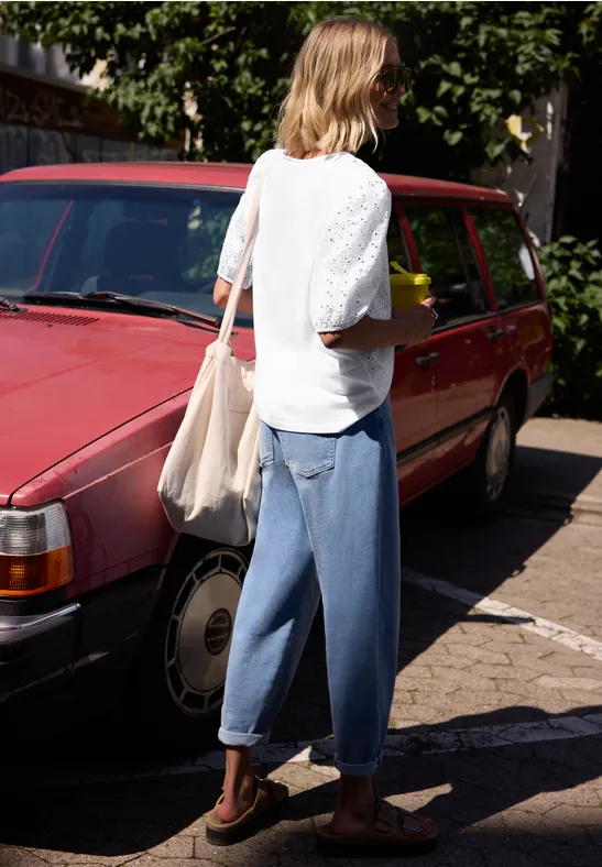 Femme en haut blanc, jean ample, sandales et sac en toile à côté d’un break rouge.