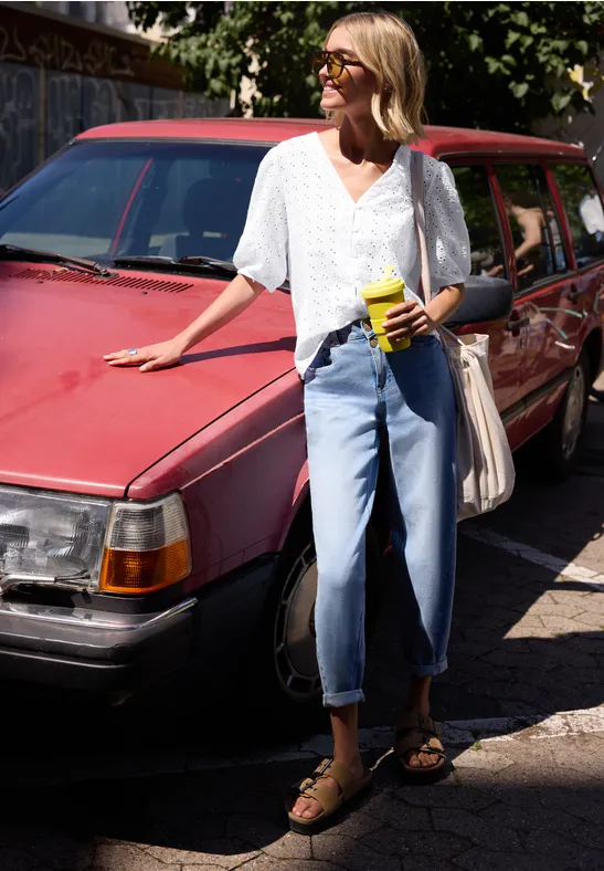 Femme en haut blanc et jean bleu clair appuyée contre une voiture rouge vintage, boisson à la main, lunettes de soleil.