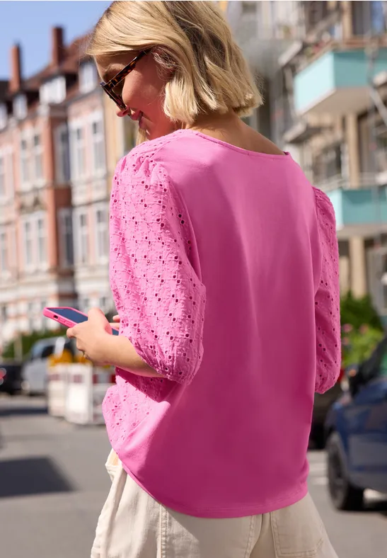 Vrouw in roze top met ajourmouwen staat op straat en bekijkt haar smartphone.
